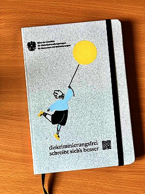 Notizblock mit der Aufschrift Diskriminierungsfrei schreibt sich's besser und der Illustration einer Person mit Beinprothese, die einen Luftballon hält
